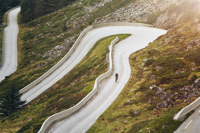 Winding mountain road with a cyclist in a scenic, hilly landscape.